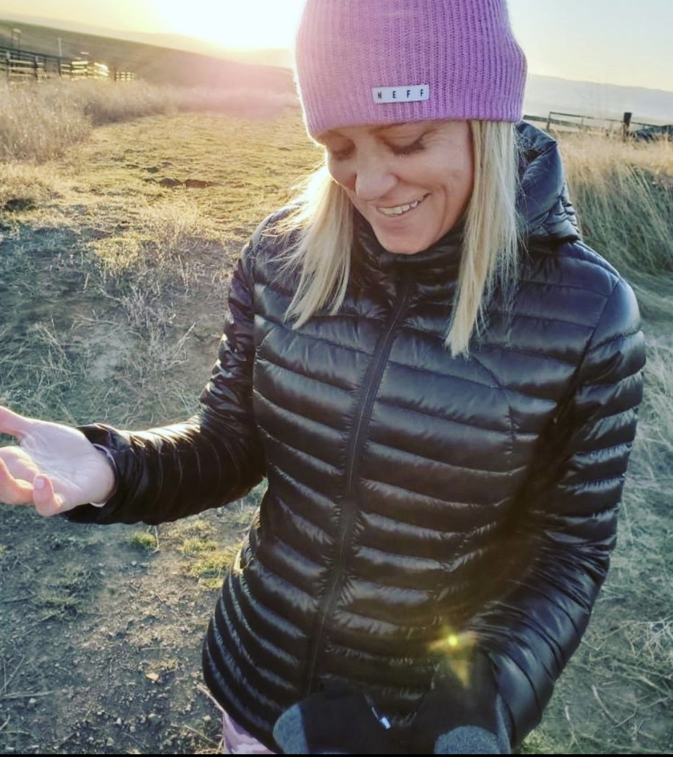 blond woman with a winter hat smiling, on a hill during the sunrise.